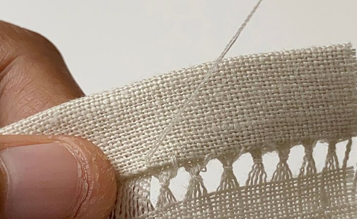 Close-up of a person performing a decorative hand-stitched hem on a cream-colored linen fabric.